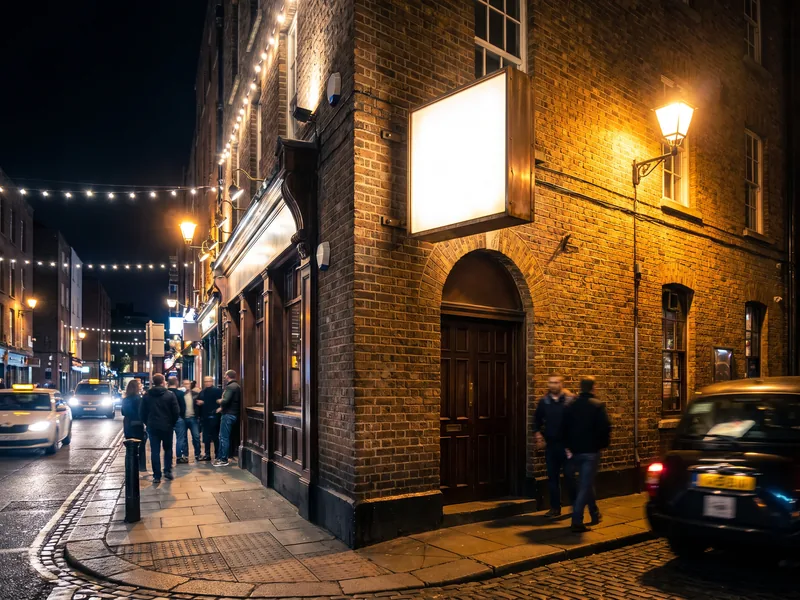 An illuminated blade sign projecting over a corner bar entrance at night