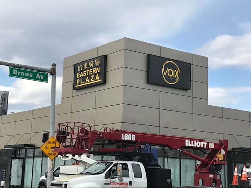 Illuminated wall cabinet reading EASTERN PLAZA in yellow on a black face with Chinese characters above it, mounted on a beige panel-clad corner building, with a VOIX cabinet on the return elevation and a boom truck parked below