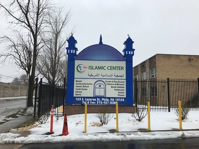 Monument sign shaped as a blue dome flanked by two crescent-topped minarets, reading ISLAMIC CENTER in English and Arabic above two columns of program listings, on a masonry base behind yellow bollards