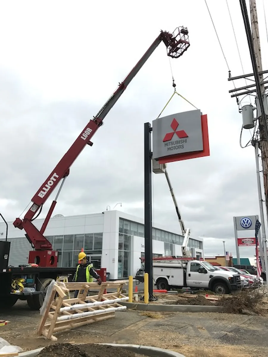 A Mitsubishi Motors sign cabinet suspended from a crane line on a two-leg sling, held just above the black steel pole it is being set onto, with a crew member standing clear on the ground