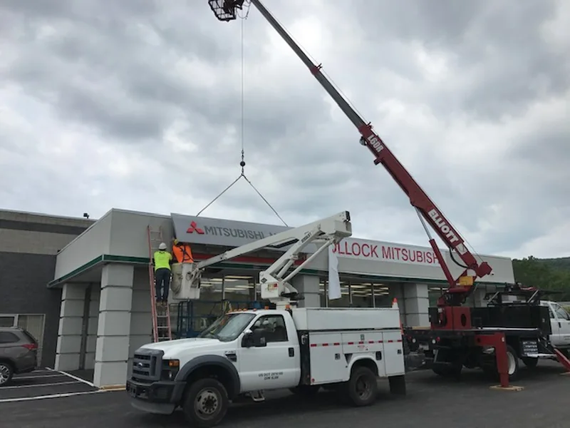 A long Mitsubishi brand fascia band suspended from a crane line above a dealership parapet, tipped down at the left end, with the dealer name band already installed below and two workers waiting on a ladder and in a bucket truck